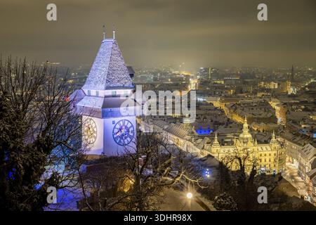 Blick auf den Uhrturm, der majestätisch gegen das Stadtbild steht, sein Gesicht beleuchtet, im Kontrast zum dunklen Himmel und schneebedeckten Bäumen, Graz, Stockfoto