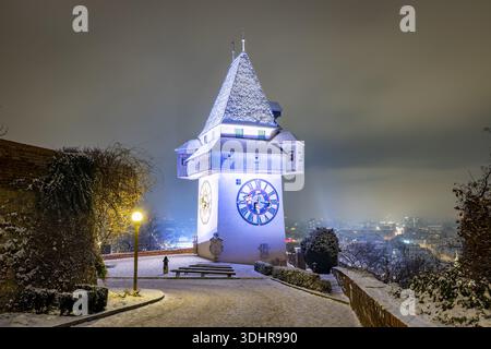 Blick auf den ikonischen Uhrturm, der majestätisch inmitten einer verschneiten Landschaft steht, dessen beleuchtetes Gesicht ein ätherisches Leuchten auf die ruhige Szene von Graz, Stockfoto
