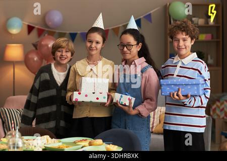 Gruppe von vier Teenagern, darunter kaukasischer Junge, kaukasischer Mädchen, asiatischer Mädchen und Junge mit möglicher gemischter Ethnie, die zusammen stehen und Geschenkboxen halten und Partyhüte tragen Stockfoto