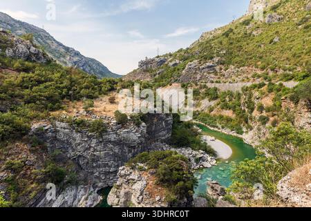 Eine dramatische Bergschlucht-Szene mit einem türkisfarbenen Moraca-Fluss, der sich durch felsige Klippen, üppige Sträucher und klaren blauen Himmel schlängelt. Raues Gelände und Grün Stockfoto