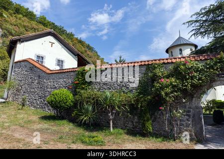 Eine bezaubernde Szene mit einer zerklüfteten Steinmauer, die in Efeu gehüllt ist, einem weißen Ferienhaus, gekachelten Dächern und üppigen Pflanzen unter einem hellen Himmel. Moraca Monas Stockfoto