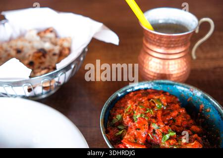 Nahaufnahme von indischem rotem Curry, serviert in einer Metallschale auf einem Holztisch, mit reichhaltiger Tomatensoße, Kräutergarnitur und traditionellem Tisch Stockfoto