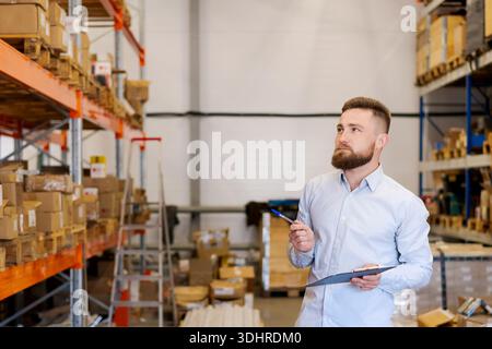 Bärtiger Vorgesetzter in einem hellen Hemd, der ein Klemmbrett hält und nachdenklich in die Regale des Lagers blickt. Konzept der professionellen Bestandsverwaltung, Audit Stockfoto