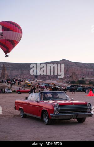 Ein klassisches Cabriolet fährt während einer Heißluftballontour bei Sonnenaufgang durch einen öffentlichen Bereich in Kappadokien, bei der die Ballons in den Himmel steigen Stockfoto