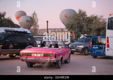 Ein klassisches Cabriolet fährt während einer Heißluftballontour bei Sonnenaufgang durch einen öffentlichen Bereich in Kappadokien, bei der die Ballons in den Himmel steigen Stockfoto