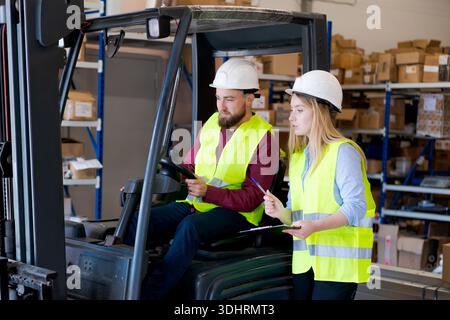 Weibliche Aufsichtsperson mit Klemmbrett, die Anweisungen für einen männlichen Gabelstaplerfahrer gibt. Logistikteam für Sicherheitsausrüstung, das Inventar und Ausrüstung in A di verwaltet Stockfoto