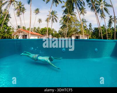 Mann schwimmt unter Wasser im Resortpool mit geteilter Aussicht auf die Wasseroberfläche oben und unten. Tropische Palmen und Hotelbungalows im Hintergrund, Stockfoto