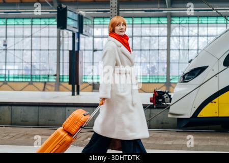 Ganzkörperfrau in weißem Kunstpelzmantel mit orangefarbenem Koffer Stockfoto