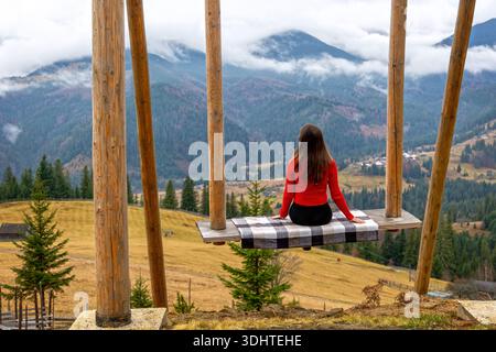 Woman on Swing mit Blick auf das malerische Bergtal. Eine Frau in einem roten Pullover sitzt auf einer Holzschaukel und genießt einen Panoramablick auf nebelige Berge und Stockfoto