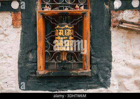 Blick auf ein Gebetsrad, das von einem rustikalen Holzfenster vor einer strukturierten Steinmauer eingerahmt wird, mit kontrastierender schwarz-weißer Farbe, Jambay Lhakhang, Stockfoto