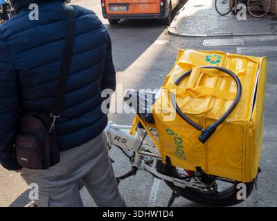 Cremona, Italien, 9. Januar 2026 Glovo-Fahrer, der neben dem Elektrofahrrad mit einem Rucksack mit gelbem Branding steht, um Lebensmittel zu liefern Stockfoto