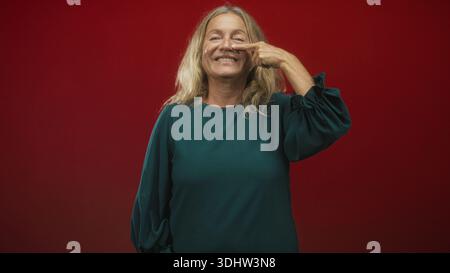 Ältere Frau mit blonden Haaren zeigt den Finger an die Nase, lächelt, trägt grüne Bluse vor rotem Studio-Hintergrund; verspieltes Selbstvertrauen. Stockfoto