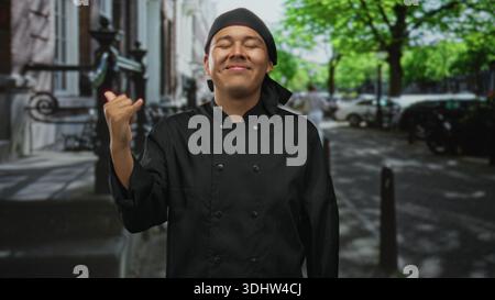 Mann Koch in schwarzer Uniform, der Koch Kuss Geste auf einer Stadtstraße mit Gesicht und Hand macht; stolz Handwerkskunst Gastfreundschaft. Stockfoto