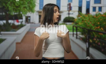 Frau zeigt Finger auf Brust auf dem Gehweg des Gebäudes, trägt weißes T-Shirt in der Nähe von Bus und Pflanzgefäßen; Überraschungsfragen. Stockfoto