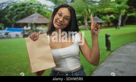Frau mit Papiertüte zeigt Zeigefinger auf dem Straßenweg des Resorts bei Poolkabanas und grünem Rasen; fröhliche Einladung. Stockfoto