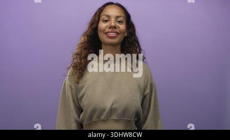 Das Gesicht der jungen Frau sieht richtig aus und trägt ein lässiges, verkürztes Sweatshirt und einen kleinen Ohrring mit Nieten im lila Studio; ruhiger Zuversicht-Optimismus. Stockfoto