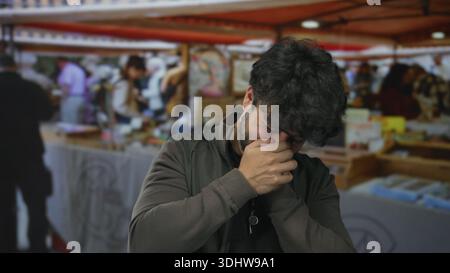 Junger Mann mit Bart lacht draußen auf einem geschäftigen Straßenmarkt mit unscharfen Menschen und bunten Verkaufsständen, die lebendige soziale Interaktion zeigen. Stockfoto