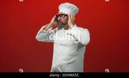 Küchenchef mittleren Alters in weißer Uniform, Kopf mit Händen berühren, Hut und Brille tragen im roten Studio; Küchenstress. Stockfoto