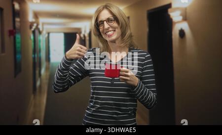 Frau, die mit der rechten Hand einen roten Becher hält und mit der linken Hand ein OK-Schild im Flur des Gebäudes macht; gemütliche Zufriedenheit. Stockfoto