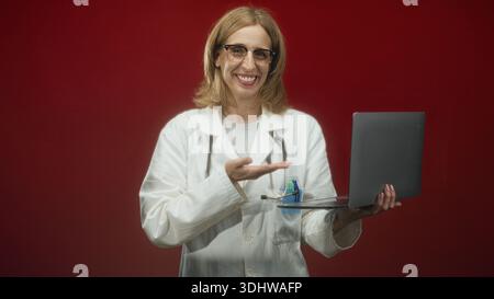 Frau Ärztin mit Laptop und Stethoskop, die im Studio mit Handbewegung vor rotem Hintergrund präsentiert, während sie lächelt und Augenkontakt herstellt; Stockfoto
