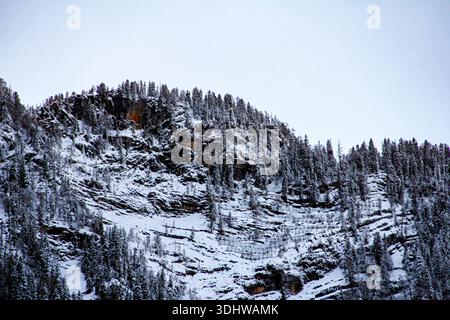 Dramatische Winterberglandschaft mit schneebedeckten felsigen Klippen und dichten immergrünen Wäldern unter einem hellen, bewölkten Winterhimmel. Stockfoto