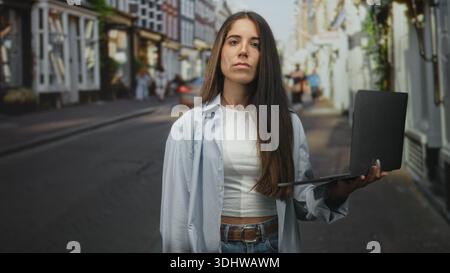 Die junge hispanische Frau zeigt mit dem Finger auf die Kamera, während sie einen Laptop hält und auf einer belebten Straße ihre Nase kneift; ekelhafte Vorsicht. Stockfoto
