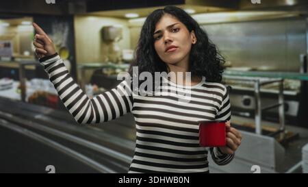 Junge brünette Frau zeigt mit dem Finger nach vorne, während sie im Küchengebäude einen roten Becher hält; Missbilligung. Stockfoto