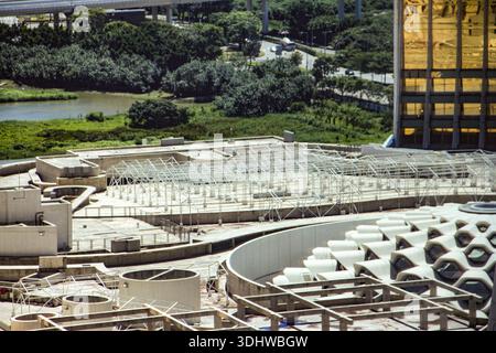 Moderne städtische Architektur mit weißen Stahlrahmen und Betonkonstruktionen vor dem Hintergrund üppiger grüner Parklandschaft und eines Goldgebäudes. Stockfoto