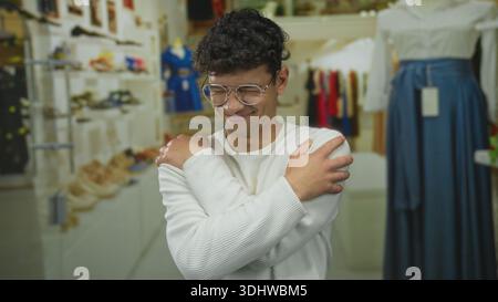 Mann mit Brille umarmt Schultern im Einzelhandelsgebäude mit verschwommenen Kleiderständern; Selbstpflegekomfort. Stockfoto