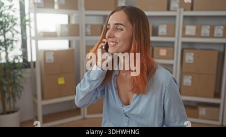 Die Frau hält das Telefon in die Ohren im Lager, zwischen gestapelten Kartonschachteln und Regalen, Sprechen und Gesten; fröhliches Unternehmertum. Stockfoto