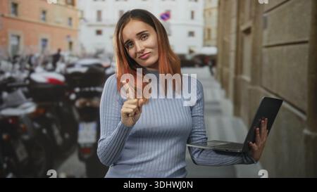 Die junge rothaarige Frau zeigt mit dem Zeigefinger und hält den Laptop, während sie einen hellblauen Pullover in einer engen Straße neben geparkten Rollern und einem Steingebäude trägt Stockfoto