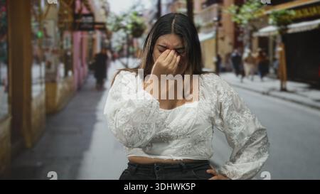 Frau mit Hand auf der Stirn für Kopfschmerzen auf der Straße, mit weißer, bestickter Bluse, zuwinkenden und reibenden Schläfen; Stress-Müdigkeit-Frustration. Stockfoto