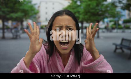 Frau im rosa Bademantel auf der Straße sieht frustriert und emotional aus, drückt im Freien unter städtischen Bäumen Wut aus und steht für emotionalen Stress und Morni Stockfoto
