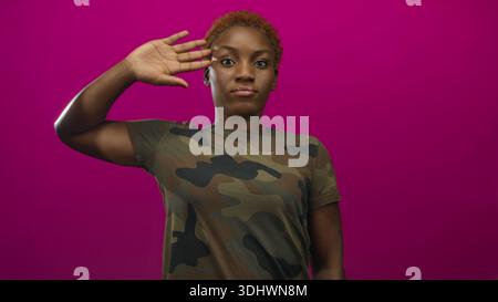 Junge afroamerikanische Frau, die mit der rechten Hand in Tarnuniform in einem leuchtend rosa Studio grüßt; Ehre. Stockfoto