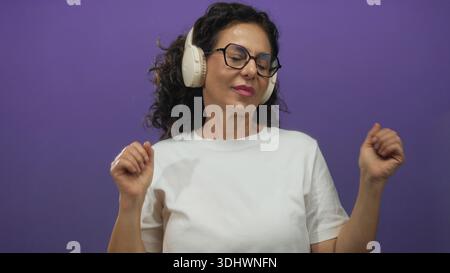 Frau genießt Musik mit Kopfhörern vor violettem Hintergrund, lächelt und tanzt in weißem Hemd, Brille, isoliert in entspanntem Casua mittleren Alters Stockfoto