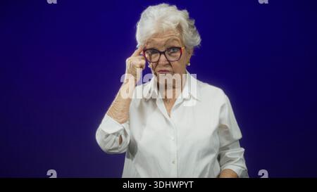 Eine ältere Frau zeigt den Zeigefinger in einem Studio auf die Kamera, trägt eine Brille, eine weiße Bluse und einen Anhänger mit einer festen Gesichtshaut; eine ernste Warnung. Stockfoto
