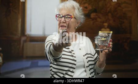 Die Seniorin zeigt den Finger nach vorne, während sie einen Miniatur-Einkaufswagen in einem kunstvollen historischen Gebäude hält; trotzt dem sparsamen Humor. Stockfoto