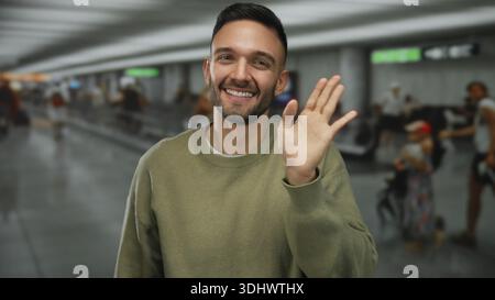 Junger Mann, der am Flughafenterminal lächelt und mit einer Welle in geschäftiger Umgebung begrüßt, die Reisen und Multikulturalität in der modernen Transitlounge mit zeigt Stockfoto