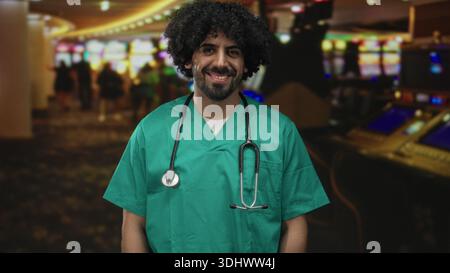 Mann Doktor in grünen Peelings mit Stethoskop lächelt neben Spielautomaten auf einem Kasino-Boden drinnen, sichtbare lockige Haare und Bart; Vertrauensdienst Stockfoto
