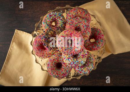 Donuts mit bunten Streuseln auf einer Glasplatte, präsentiert auf einem Holztisch mit gelbem Tuch Stockfoto