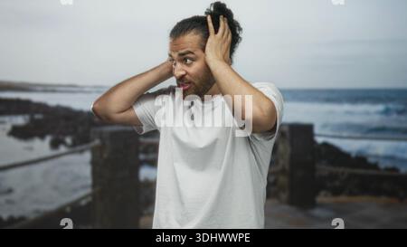 Der Mann bedeckt die Ohren mit Händen am Straßenpier am Meer, hält den Kopf und grimmt; überwältigte Frustration. Stockfoto