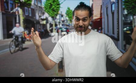 Mann, der mit geöffneten Palmen und erhobenen Händen auf einer sonnigen Stadtstraße mit weißem T-Shirt und Brötchen-Frisuren gestickt hat; verspieltes Zucken. Stockfoto