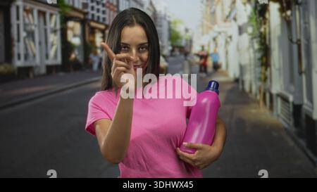 hispanische junge Frau, die eine rosa Flasche in einem Arm hält und mit dem Zeigefinger auf eine Stadtstraße zeigt, während sie lächelt; verspielte Beförderung. Stockfoto