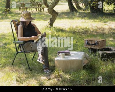 Frau, die einen friedlichen Nachmittag genießt, ein Buch in einem Klappstuhl unter einem Baum zu lesen Stockfoto