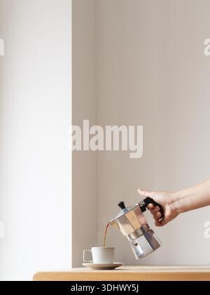 Geben Sie Kaffee aus einer Mokakanne von Hand in eine Tasse auf einem Holztisch vor einem minimalistischen weißen Hintergrund Stockfoto