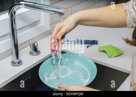 Frauenhände schrubben eine türkisfarbene Schüssel unter einem laufenden Chromhahn mit Seifenschaum in einem modernen, weißen Küchenbecken Stockfoto