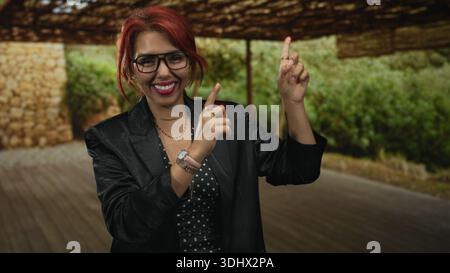Frau zeigt den Zeigefinger nach oben auf die Veranda, lächelt mit Brillen und mehrschichtigen Ketten; Freude am Vertrauen. Stockfoto