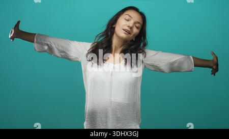 Frau, die die Arme weit ausdehnt, die Augen geschlossen und ein leichtes Lächeln trägt, weiße Bluse in einem tealfarbenen Studio-Setting; ruhige Reflexion. Stockfoto