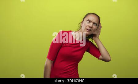 Frau, die mit der Hand das Ohr schröpft, den Kopf neigt, sich vor die lindgrüne Studiowand lenkt und dabei rotes Hemd trägt; Neugier-Aufmerksamkeit. Stockfoto