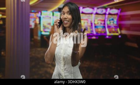 Die junge thailändische Frau, die ein Smartphone hält, hebt die Hand auf, um in der Nähe farbenfroher Spielautomaten im Gebäude anzuhalten; fröhlicher Gruß. Stockfoto
